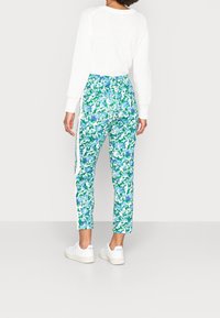 Osoba oblečená v bílém pleteném svetru a květinových kalhotách s bílým bočním pruhem, doplněná bílými teniskami, stojí před bílým pozadím.