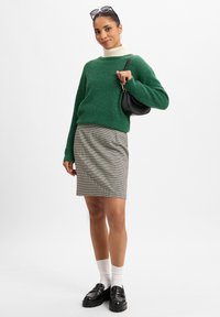 Grüner gestrickter Pullover, weißer Rollkragenpullover darunter, grauer Karorock, schwarze Umhängetasche, weiße Socken und schwarze Slipper.