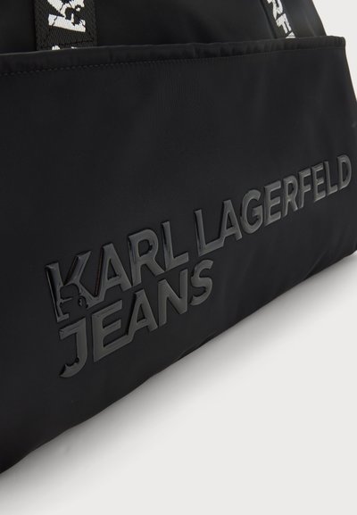Едър план на черна чанта Karl Lagerfeld Jeans с релефни букви на логото и черни дръжки с бели тъкани детайли.
