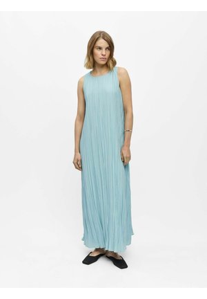 Woman with shoulder-length blonde hair wearing a sleeveless, light blue pleated maxi dress and black flat shoes, standing against a plain white background.