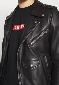 Zbliżenie na osobę w czarnej skórzanej kurtce motocyklowej, noszącą czarną koszulkę z czerwonym i białym logo "LEVI'S".