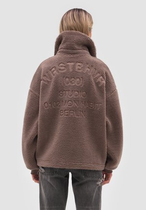 Person iført en brun fleecejakke med høj krave og præget tekst på ryggen, kombineret med mørke, slidte jeans, stående mod en grå baggrund.