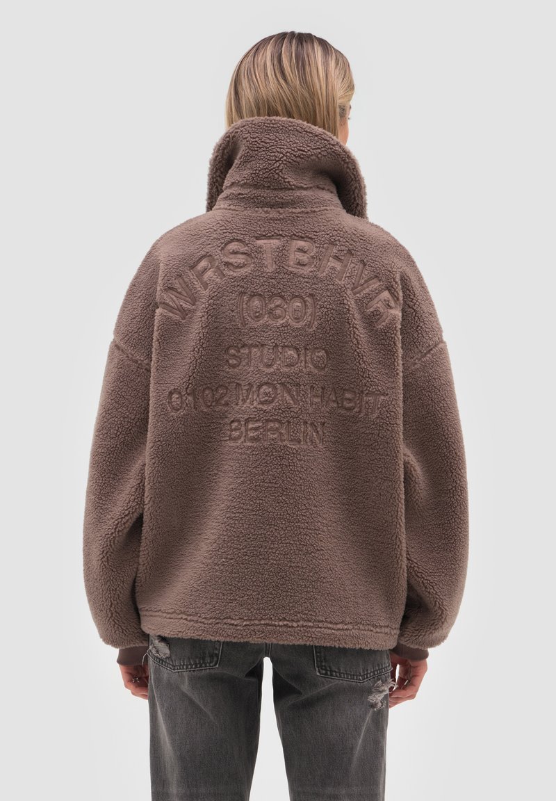 Henkilö, jolla on päällään ruskea fleece-takki korkealla kauluksella ja selässä kohokuvioitu teksti, yhdistettynä tummiin revittyihin farkkuihin, seisoo harmaata taustaa vasten.