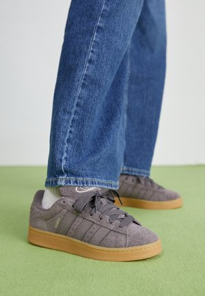 Personne portant des baskets Adidas en daim gris avec semelles en gomme, chaussettes blanches et jean bleu en denim, debout sur un tapis vert.
