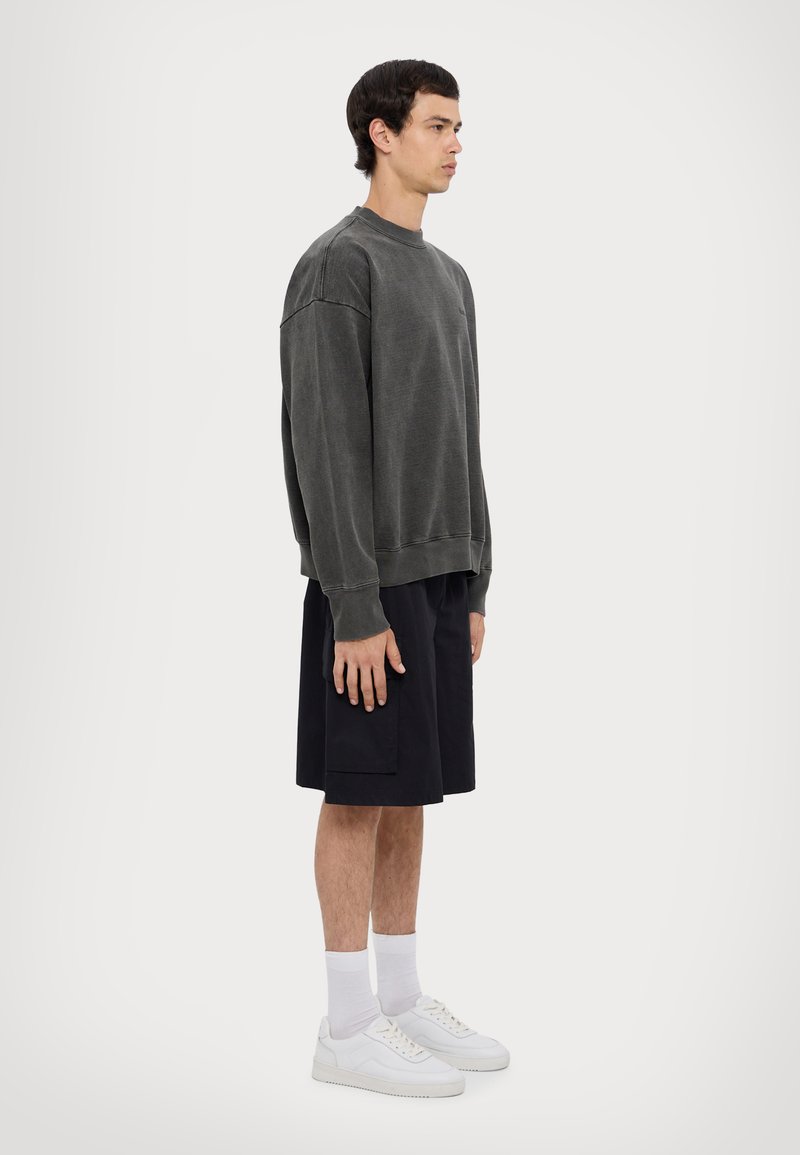 Grauer übergroßer Pullover mit gerippten Bündchen, kombiniert mit dunklen Shorts und weißen Sneakers. Das Model steht im Profil vor einem schlichten Hintergrund.
