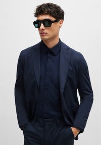 Navy suit with a slim fit, paired with a navy button-up shirt. Black square sunglasses with dark lenses. Smooth fabric texture throughout.