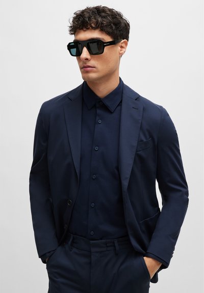 Costume marine ajusté, associé à une chemise marine à boutons. Lunettes de soleil carrées noires avec des verres foncés. Texture de tissu lisse sur l'ensemble.
