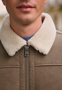 Braune Lederjacke mit einem cremefarbenen Kunstfellkragen, vorne mit Reißverschluss und strukturiertem Stoff. Der Kragen hat eine kontrastierende, weiche Oberfläche.