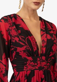 Vestito rosso a fiori con scollatura a V profonda e tessuto nero. Presenta una vita arricciata e maniche lunghe e trasparenti.