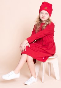 Młoda dziewczynka z kręconymi blond włosami, ubrana w czerwoną sukienkę, czerwoną czapkę beanie i białe trampki, siedząca na jasnym drewnianym stołku na beżowym tle.