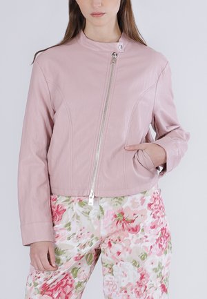 Kvinde iført en lyserød lynlåstrøje med den ene hånd i lommen, kombineret med hvide bukser med et pink og grønt blomsterprint.