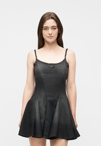 Νεαρή γυναίκα με ίσια καστανά μαλλιά που φοράει μαύρο μαν sleeveless denim φόρεμα με φουσκωτή φούστα, στέκεται μπροστά σε απλό φόντο.