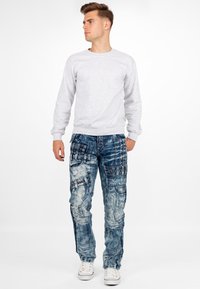 Felpa grigia chiara, jeans blu strappati con molte tasche, borchie e un lavaggio sbiadito; indossata con sneaker bianche.