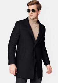 Czarny wełniany płaszcz z wyłamiem, dopasowany krój i dwoma bocznymi kieszeniami. Połączony z beżowym golfem i zielonymi okularami przeciwsłonecznymi.