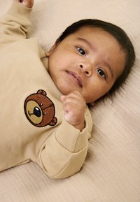 Smėlio spalvos kūdikių bodis su rudu meškiuku išsiuvinėtu ant krūtinės. Minkšta medžiaga, riboti rankogaliai ir plokščios siūlės šviesiai smėlio spalvos fone.