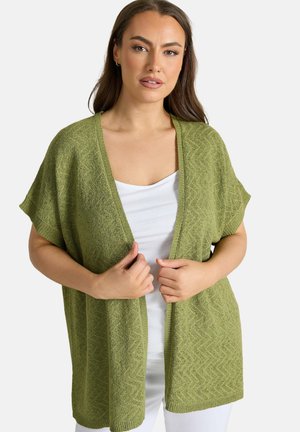 Kvinde iført en grøn åben-front strikket cardigan over en hvid top, som holder cardiganens kanter med begge hænder.
