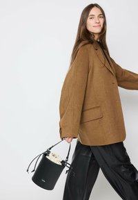 Brown oversized blazer with a notched collar, paired with black leather pants. The model holds a round black handbag with a drawstring closure.