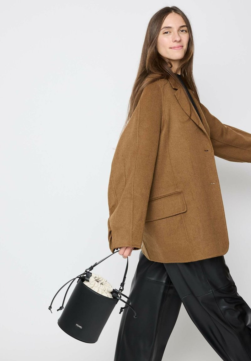 Brown oversized blazer with a notched collar, paired with black leather pants. The model holds a round black handbag with a drawstring closure.