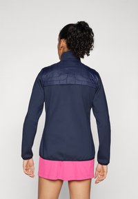 Tummansininen takki, jossa on tilkutettu, vaaleansininen olkapala; pitkät hihat, korkea kaulus; yhdistetty vaaleanpunaiseen hameeseen, minimalistiinen muotoilu.