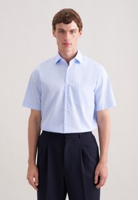 Jasnoniebieska koszula na krótki rękaw z kieszenią na piersi, noszona z ciemnymi, dopasowanymi spodniami. Gładki materiał i tradycyjny kołnierzyk.