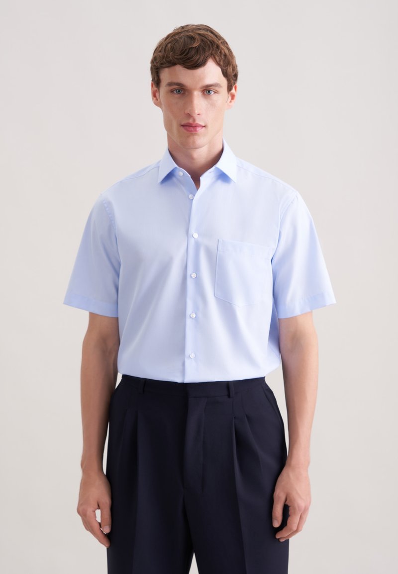 Jasnoniebieska koszula na krótki rękaw z kieszenią na piersi, noszona z ciemnymi, dopasowanymi spodniami. Gładki materiał i tradycyjny kołnierzyk.