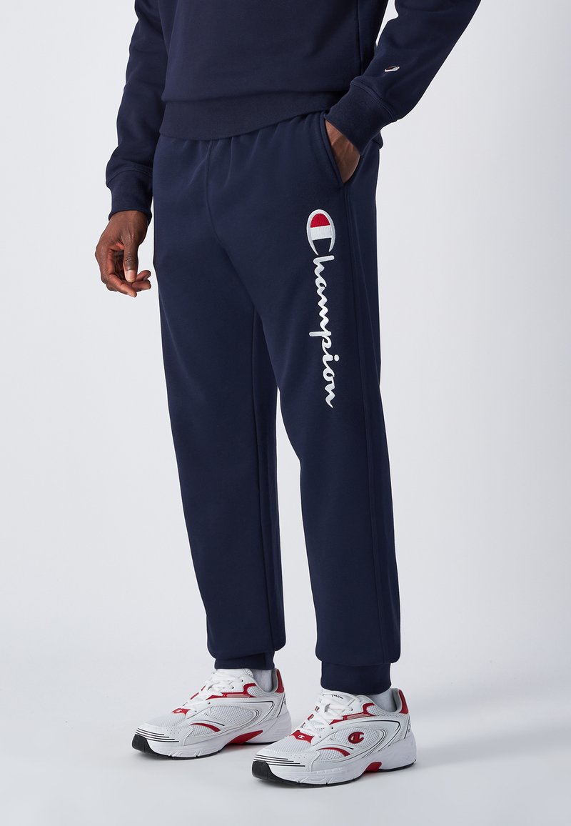 Pantaloni della tuta blu marina realizzati in tessuto morbido, con un logo bianco "Champion" sulla gamba sinistra e tasche laterali. Abbinati a scarpe da ginnastica bianche e rosse.