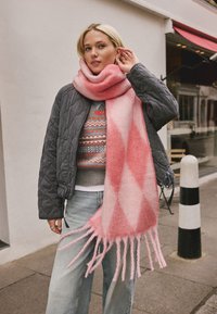 Long pink and white checkered scarf with fringing, worn with a gray quilted jacket and patterned sweater, set against an urban backdrop.