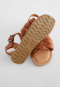Brun lädersandal med flera korsade remmar, justerbar spänne och en texturerad ljusbrun gummisula med ett vågigt mönster.