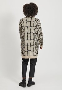 Lång, beige cardigans med svart rutmönster, tillverkad av mjukt, texturerat tyg. Har breda ärmar och en rundad nederkant. Svarta byxor och ankelstövlar.