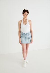 Body alb cu guler halter, asortat cu o fustă mini din denim bleu deschis, având două buzunare frontale și un fermoar, purtat cu adidași albi.
