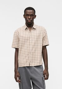 Πουκάμισο με κοντά μανίκια σε σχεδιασμό καρό ανοιχτού καφέ χρώματος, με κουμπιά στο μπροστινό μέρος, μυτερό γιακά και άνετη εφαρμογή. Το ύφασμα φαίνεται υφασμένο.