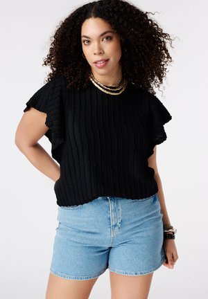 Vrouw met krullend haar die een zwart gestructureerd kortermig topje draagt, lichtblauwe denim shorts, gouden ketting en armbanden, staand tegen een witte achtergrond.