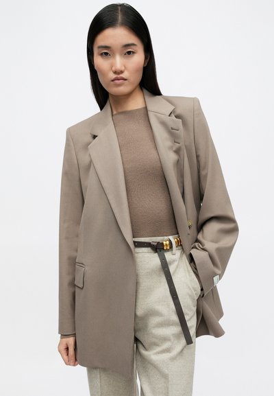 Μπεζ καλοραμμένο σακάκι με δομημένο σχέδιο, μονό κουμπί και πλαγιές τσέπες, συνδυασμένο με καφέ εφαρμοστό τοπ και μπεζ παντελόνι, τονισμένο από μια καφέ ζώνη.