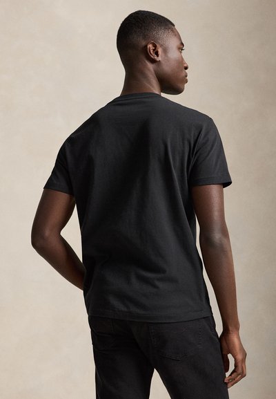 Moški, prikazan od zadaj, oblečen v enostavno črno majico s kratkimi rokavi in črne hlače na nevtralnem ozadju.