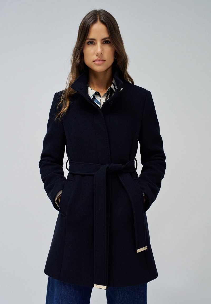 Manteau en laine bleu marine avec une taille ceinturée, un col haut et des poches latérales. Présente des accents en métal argenté et une coupe ajustée, atteignant le milieu des cuisses.