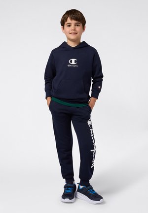 Ragazzo in piedi con le mani in tasca, indossa un felpa Champion blu navy e pantaloni sportivi, insieme a scarpe da ginnastica blu, su sfondo bianco.