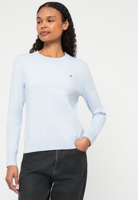 Gaiši zils adīts džemperis ar apaļu kakla izgriezumu un ribotām piedurknēm, ar mazu logotipu kreisajā krūtī, apvienots ar tumši pelēkiem biksēm.