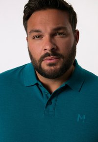 Teal polotröja med krage, tre knappar och ett texturerat tyg. Har en liten, präglad logotyp på vänster bröst.