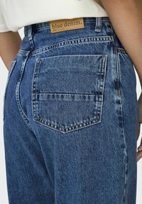 Modré džíny z denimu s vysokým pasem, obsahující hnědou koženou nášivku s nápisem "modrý denim" a dvě zadní kapsy s jemnými stehovými detaily.