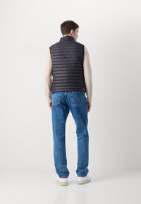 Gilet nero trapuntato con colletto alto, indossato su un maglione beige chiaro e abbinato a jeans blu. Sneakers bianche completano l'outfit.
