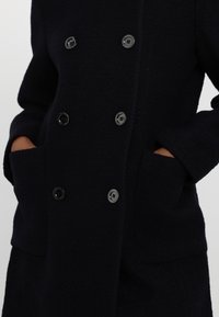 Manteau noir à double boutonnage texturé avec six boutons, deux grandes poches avant ; mains partiellement dans les poches, sur un fond clair uni.