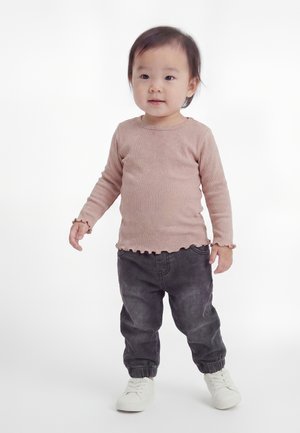 Copil mic stând, purtând un tricou roz cu mâneci lungi, pantaloni gri închis și adidași albi, pe un fundal alb simplu.