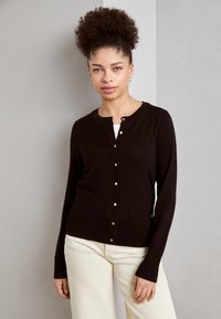 Bruine gebreide cardigan met een ronde hals, knoopsluiting aan de voorkant en lange mouwen. Soepele textuur en ribgebreide manchetten, gecombineerd met beige broek.