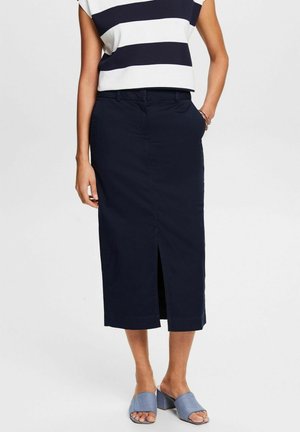 Mujer que lleva una falda midi azul marino con una abertura frontal, una blusa sin mangas a rayas blancas y azul marino, y sandalias de tacón ancho azul con punta abierta.