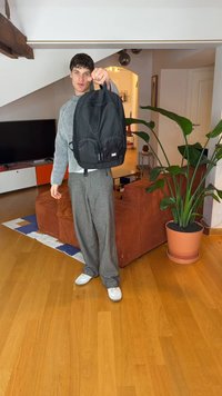 Juoda kuprinė su dviem priekinėmis kišenėmis, reguliuojamomis dirželiais ir viršutine rankena; pagaminta iš tvarios medžiagos, pateikta jaukioje vidaus aplinkoje.