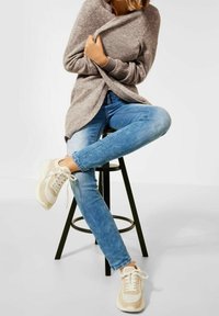 Osoba sedící na černém stoličce, v lehce modrých džínách, béžových a bílých teniskách a pohodlném šedém svetru, který si drží.