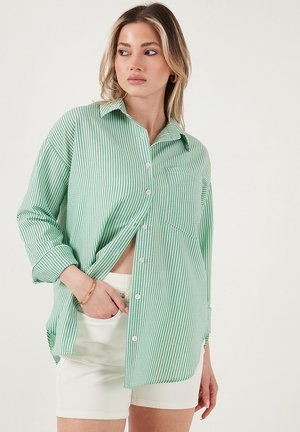 Modelka oblečená vo veľkej, zeleno-biele pruhovanej košeli, čiastočne rozopnutej, v kombinácii s bielymi šortkami, díva sa do strany.