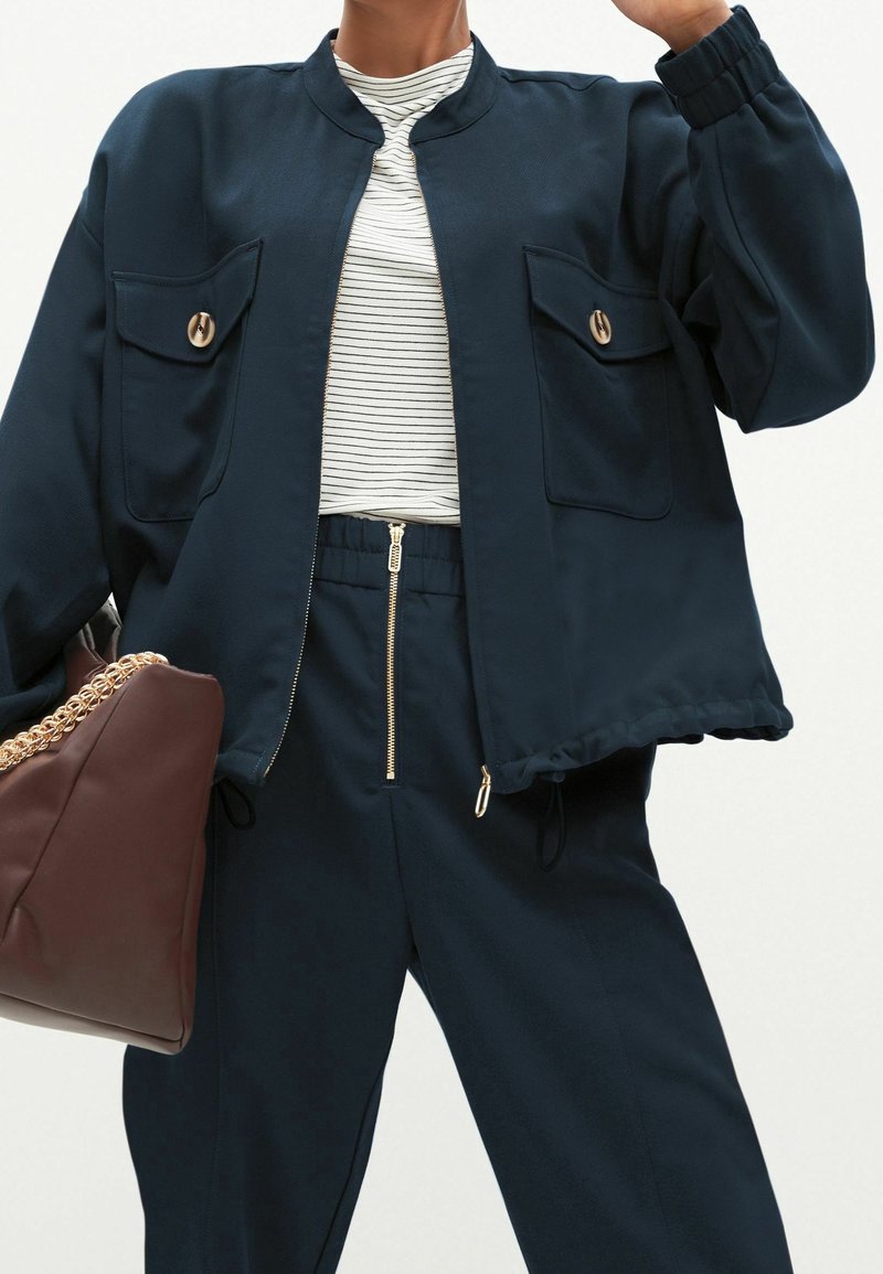 Personne portant une veste zippée bleu marine avec de grandes poches à boutons, un pantalon assorti avec une fermeture éclair frontale, une chemise rayée, et portant un sac marron avec une chaîne dorée.