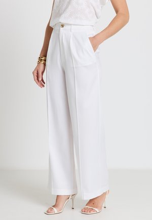 Woman wearing high-waisted wide-leg white trousers with gold button, paired with white strappy heels and gold bracelets.