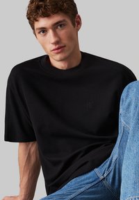T-shirt nera a maniche corte con collo rotondo e tessuto testurizzato. Presenta un logo goffrato discreto sul petto. Abbinata a jeans in denim azzurro chiaro.